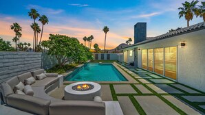 A modern outdoor pool area with a built-in sofa and a fire pit, surrounded by palm trees and a clear sky. - Luxury 3 bed 2 bath pool spa home with 29 day minimum (Palm Desert)