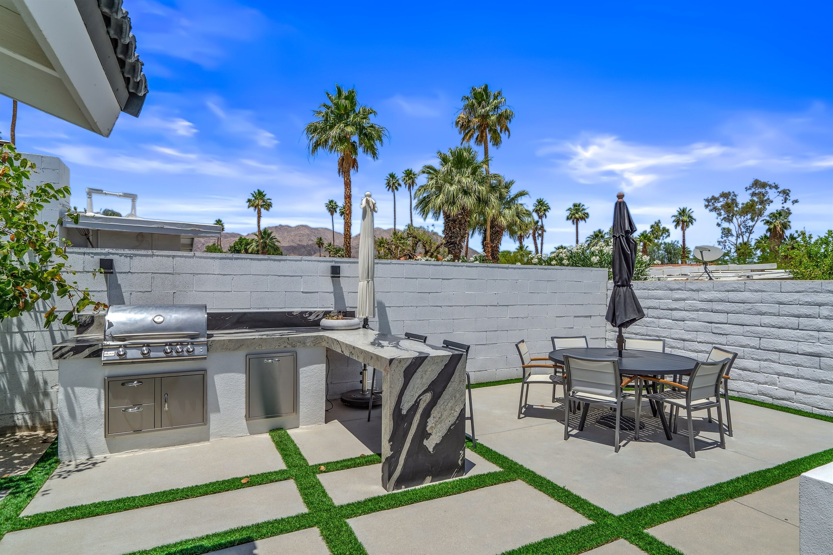 An outdoor kitchen with a grill, dining table, and chairs, surrounded by a white wall and palm trees.