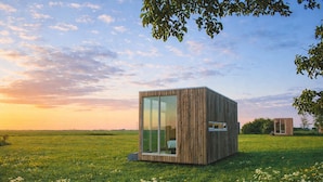 Eine moderne Holzhütte mit Glastür, eingebettet in eine grüne Wiese, im Hintergrund ein Sonnenuntergang.