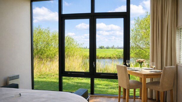Ein Schlafzimmer mit großem Fenster, einem Bett, einem Tisch mit Stühlen und Blick auf einen See und Grünflächen.