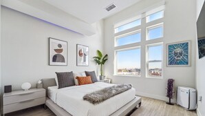 Un dormitorio moderno con una cama grande, una mesita de noche, una planta en maceta y vistas a la ciudad a través de amplias ventanas.