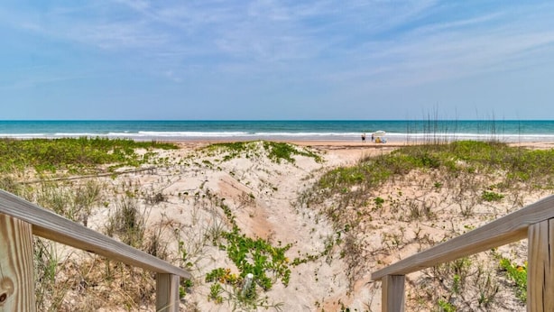 Vue sur la plage depuis une terrasse en bois, avec un paysage sablonneux et une végétation clairsemée.