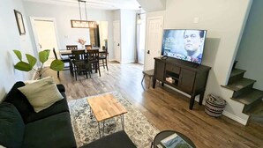 Un salon avec un canapé noir, une table basse en bois et un téléviseur posé sur un meuble. On aperçoit à l’arrière-plan un coin repas avec une table et des chaises.