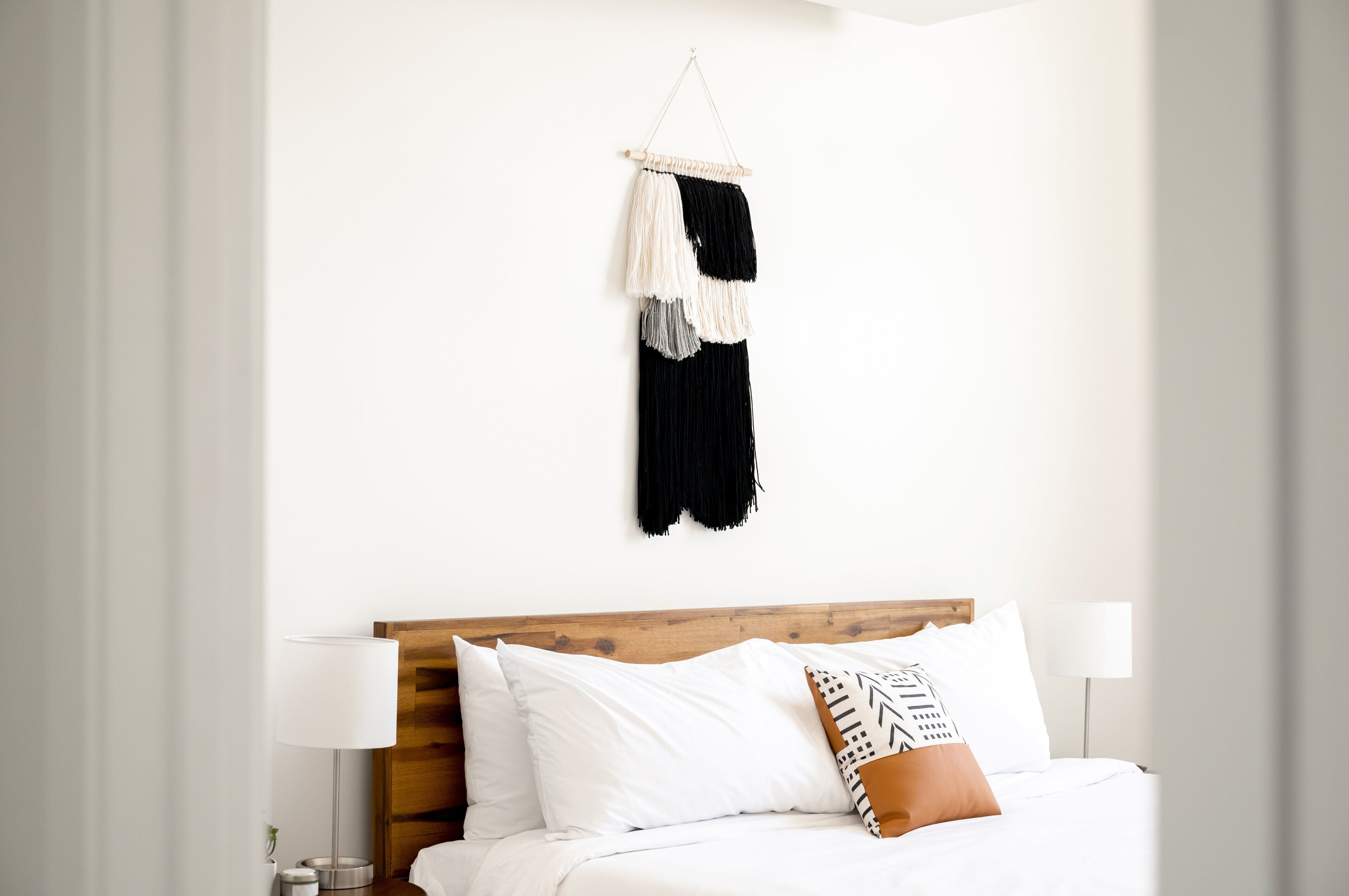 A neatly made bed with white linens and a wooden headboard, a hanging macrame wall decoration, and a bedside lamp.