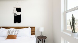 A hotel room with a wooden headboard, white bedding, a round side table with a lamp, a macrame wall hanging, and a view of a building through the window. - 2880 U505 Skytop Perspective at Espadin (Denver)