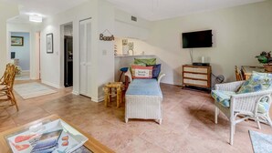 Una sala de estar con ventilador de techo, televisor, mobiliario de mimbre y una cocina al fondo.