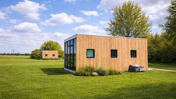 Una cabana de fusta moderna amb grans finestres, situada en un camp verd amb un cel serè.