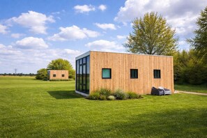Una cabaña moderna de madera con ventanas amplias, ubicada en un campo cubierto de hierba bajo un cielo despejado.