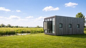 Ein modernes, minimalistisches Gebäude mit großen Fenstern und einer Holzfassade, gelegen in einem üppigen Grünland mit einem kleinen Gewässer in der Nähe.