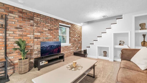 A living room with a brick wall, a flat-screen TV, a coffee table, a sofa, and a staircase with white railings. - 2 min to AUD | Private Getaway w/ Firepit (Kitchener)