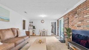 A living room with a brown leather sofa, a coffee table, a TV on a brick wall, and a dining area in the background. - 2 min to AUD | Private Getaway w/ Firepit (Kitchener)