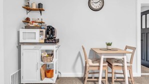 A kitchen with a microwave, coffee maker, and shelves with kitchen items. There is a clock on the wall and a table with chairs. - 2 min to AUD | Private Getaway w/ Firepit (Kitchener)