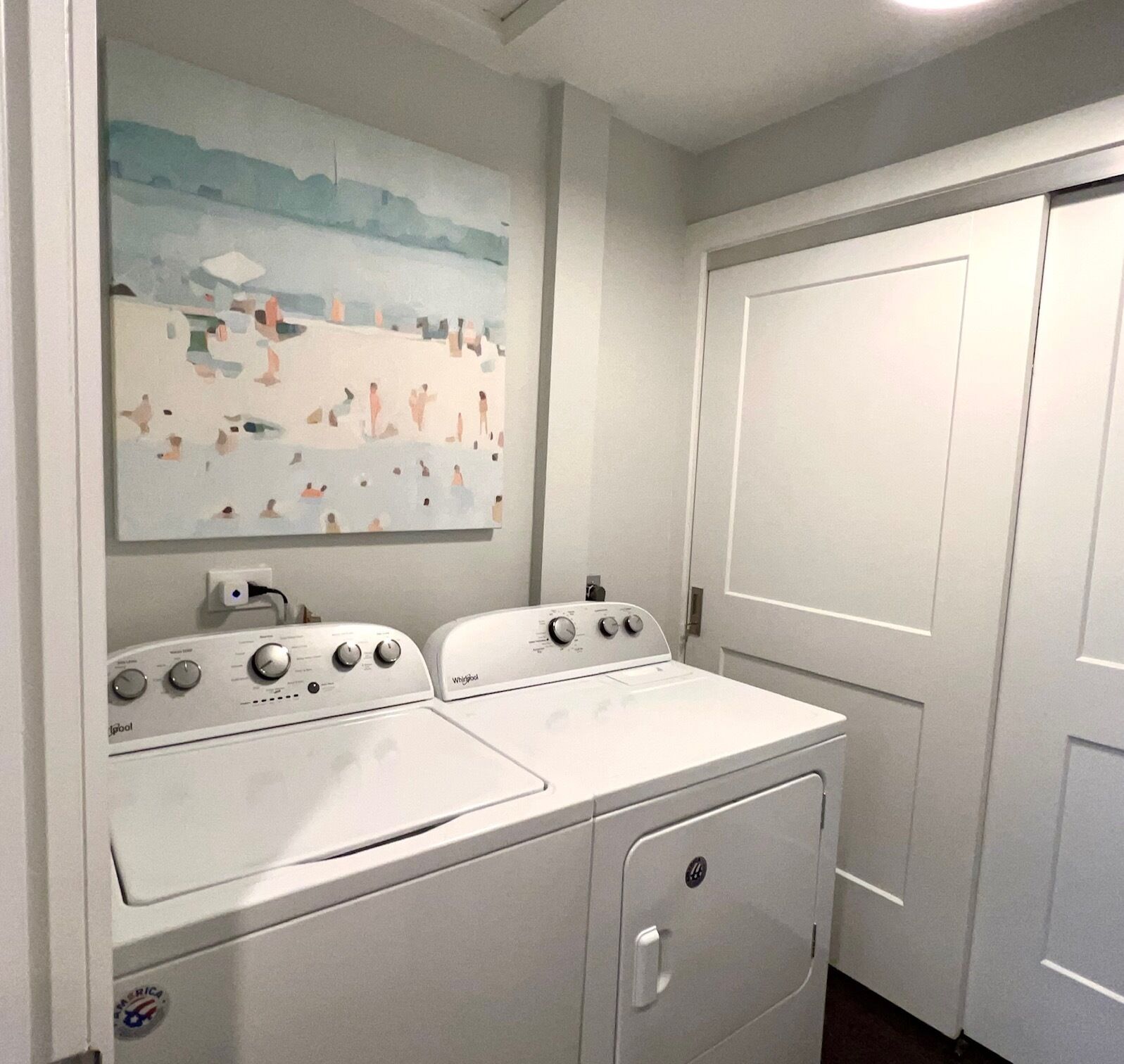 Un cuarto de lavado con lavadora y secadora Whirlpool blancas, un cuadro de una playa y una puerta cerrada.