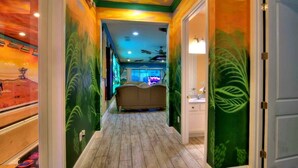 Un couloir avec un plancher en bois, des murs verts ornés de motifs de feuilles, un ventilateur de plafond et, au loin, un lit.