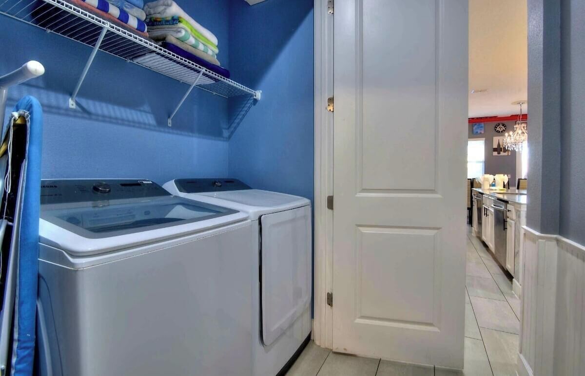A laundry room with two washing machines, a drying rack, and a door leading to a kitchen area.