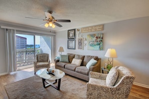 A living room with a brown sofa, patterned armchairs, a coffee table, and a ceiling fan. There are framed pictures and a sign on the wall. - 243 El Matador (Fort Walton Beach)