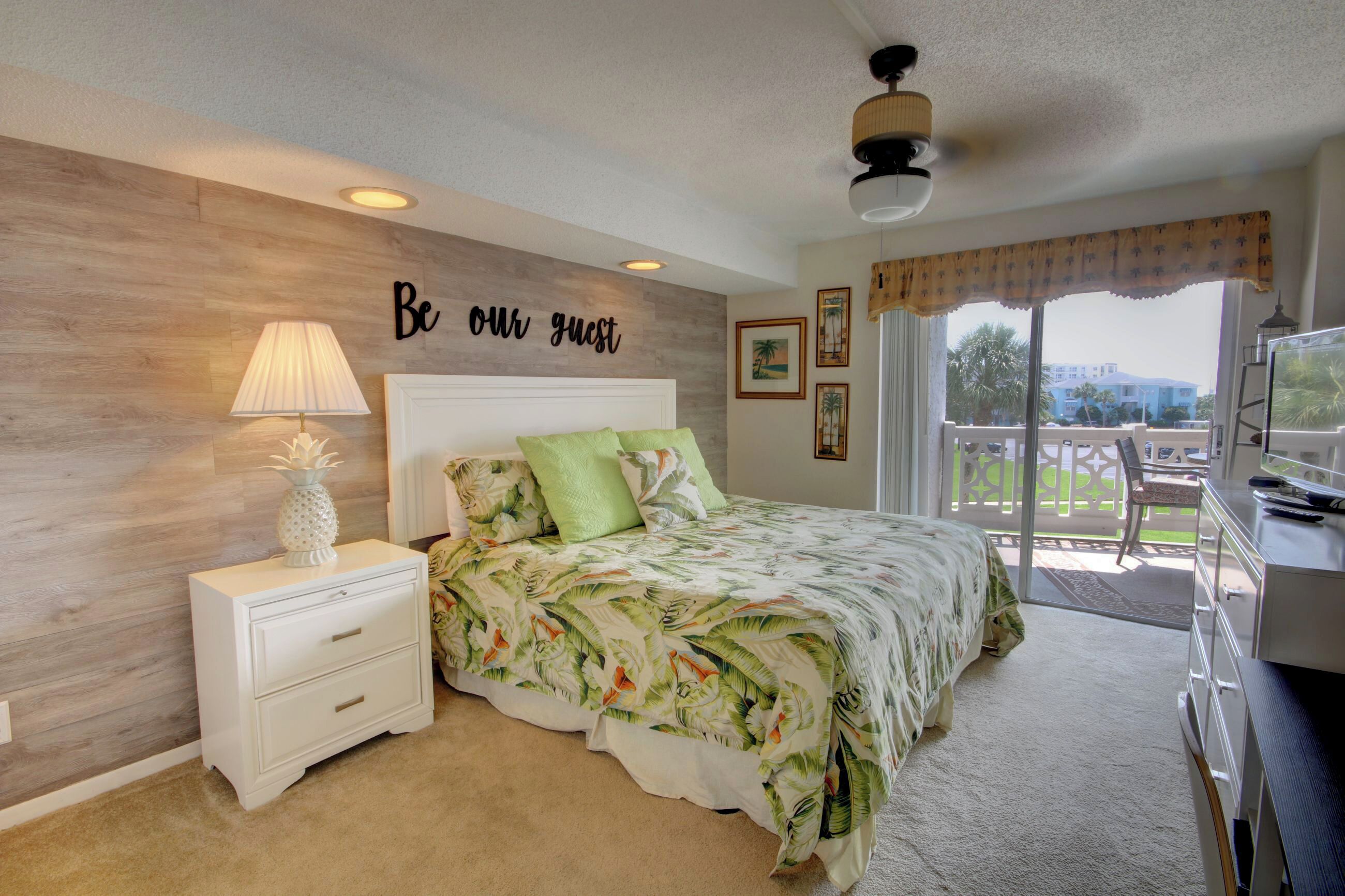 Un dormitorio con cama, mesita de noche, lámpara, ventilador de techo, ventana con vistas y un cartel que dice 'Be our guest'.