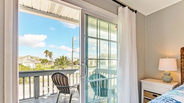 A room with a sliding glass door leading to a balcony with a chair and a table. There is a bed with a blue and white patterned bedspread and a lamp on a bedside table. - 2810 Ohio Avenue #1 (Fernandina Beach)