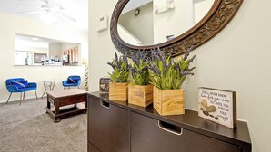 A room with a dark wooden dresser, a round mirror with an ornate frame, and three wooden boxes with lavender plants. There are two blue chairs and a small table in the background. - 2663 Andros Lane, Kissimmee, 3 BED (Kissimmee)