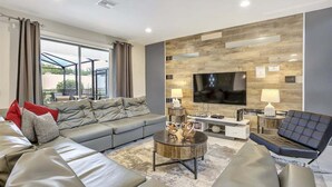 A modern living room with a grey sofa, a black armchair, a coffee table, and a flat-screen TV mounted on a wooden wall. - 2440-5-td-perfect Villa at Windsor (Kissimmee)