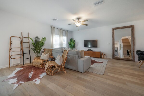 A modern living room with a grey sofa, wicker chairs, a wooden ladder, a ceiling fan, a flat-screen TV, and a large mirror. - 2405 E 19th Ave (Tampa)