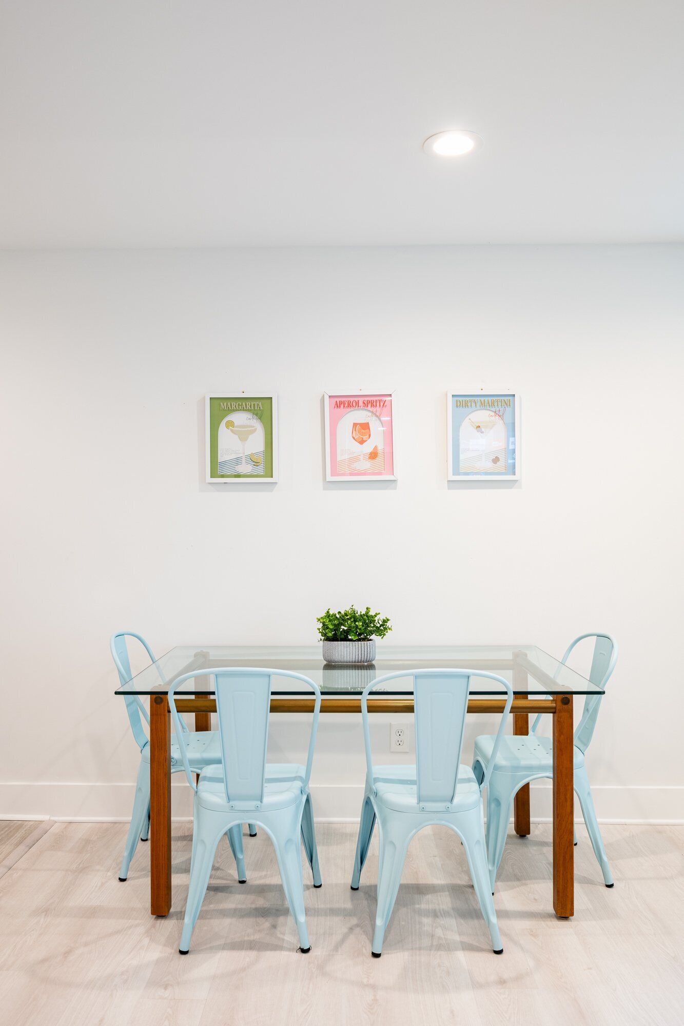 Un coin repas avec une table en verre et quatre chaises bleu clair, une plante en pot sur la table et trois tableaux encadrés au mur.