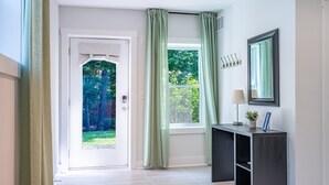 Een gang met een deur naar een tuin, een raam met gordijnen, een donkere console tafel met een lamp en een spiegel aan de muur.