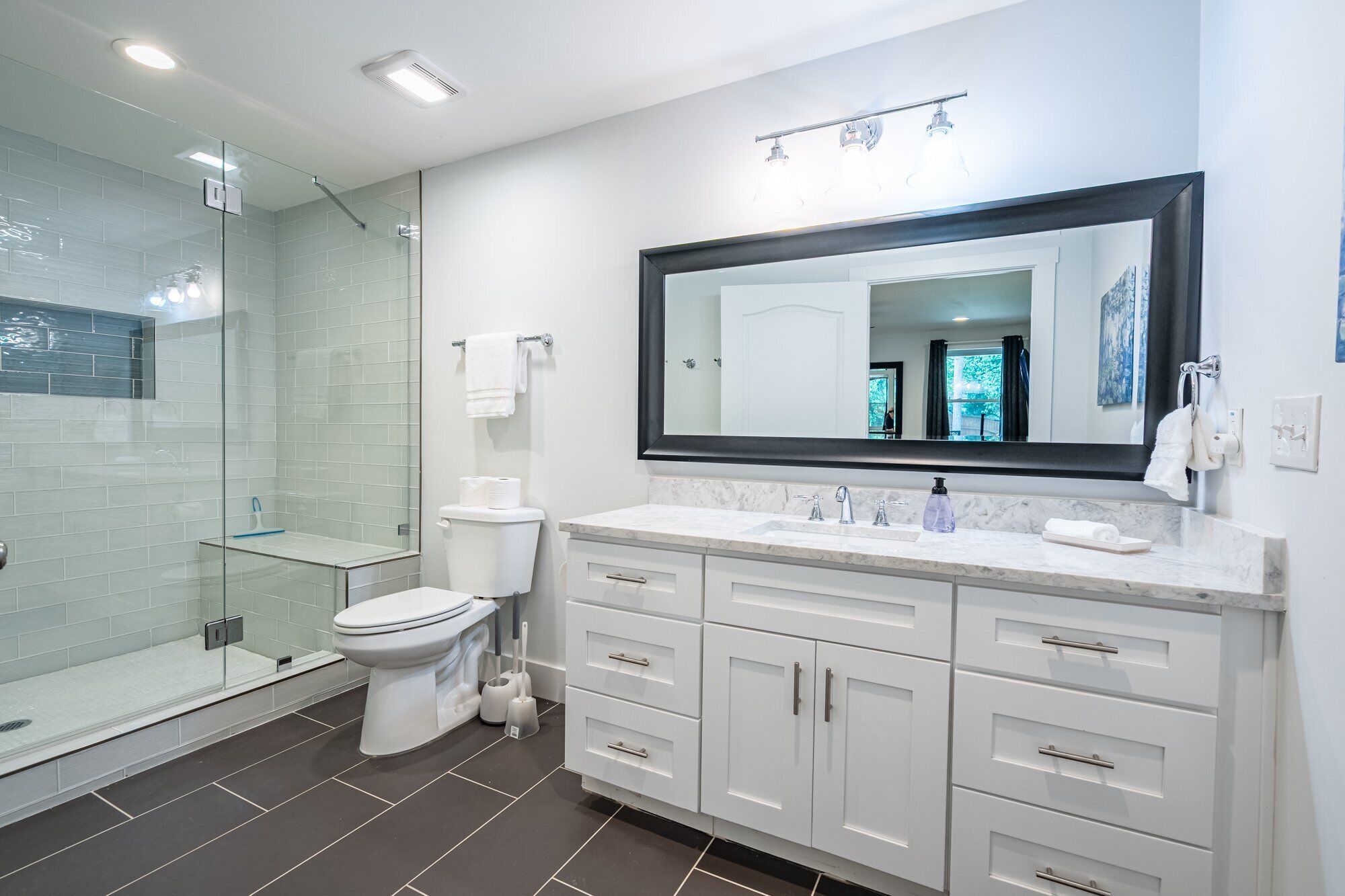 Une salle de bain moderne avec une douche à l’italienne en verre, des accessoires blancs et un grand miroir au-dessus du lavabo.