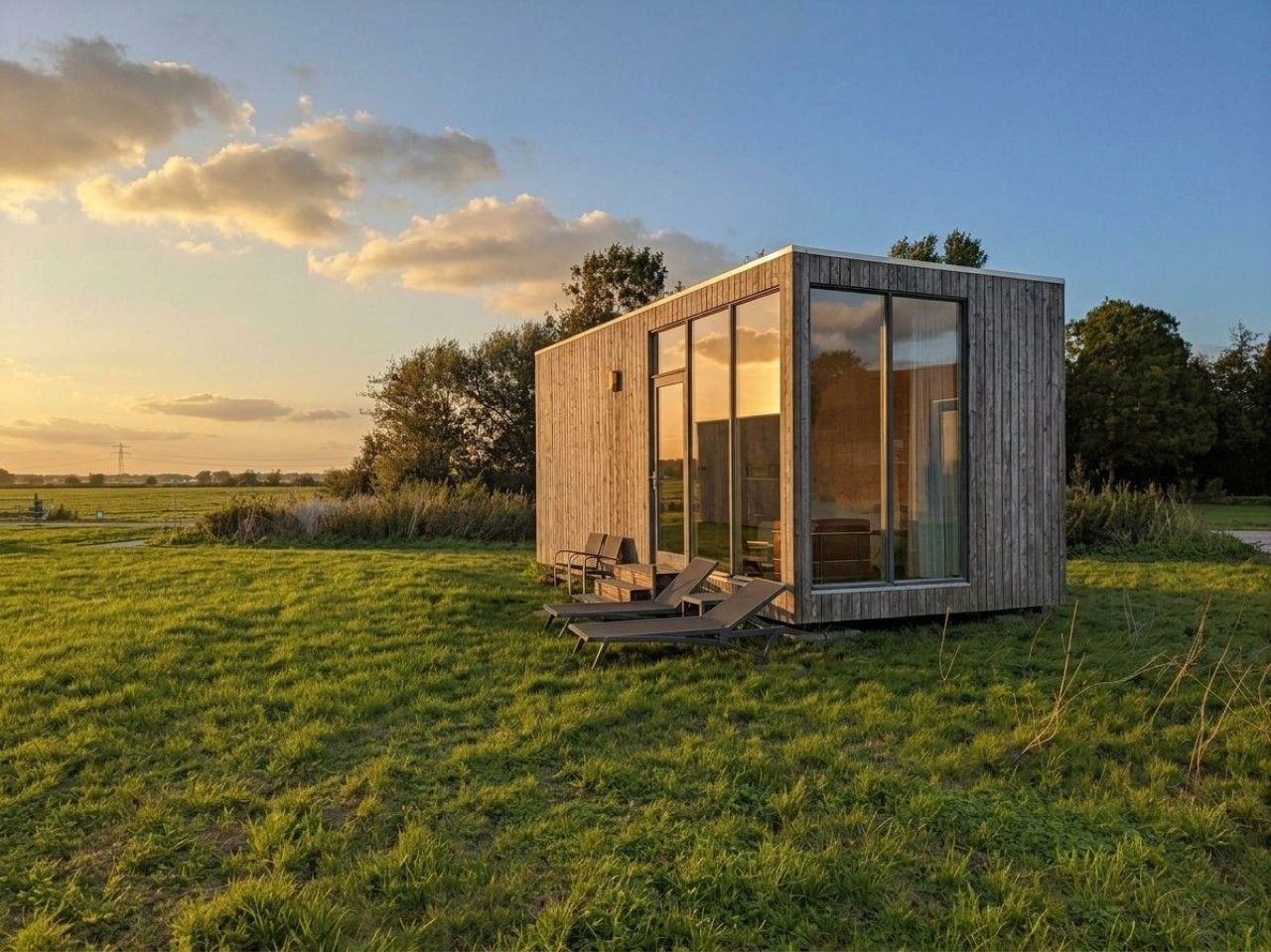 Eine moderne, eingeschossige Holzblockhütte mit großen Glasfenstern, gelegen auf einer grünen Wiese unter blauem Himmel.