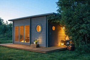 Une cabane moderne dotée de grandes fenêtres, d’une terrasse et d’une porte ronde.