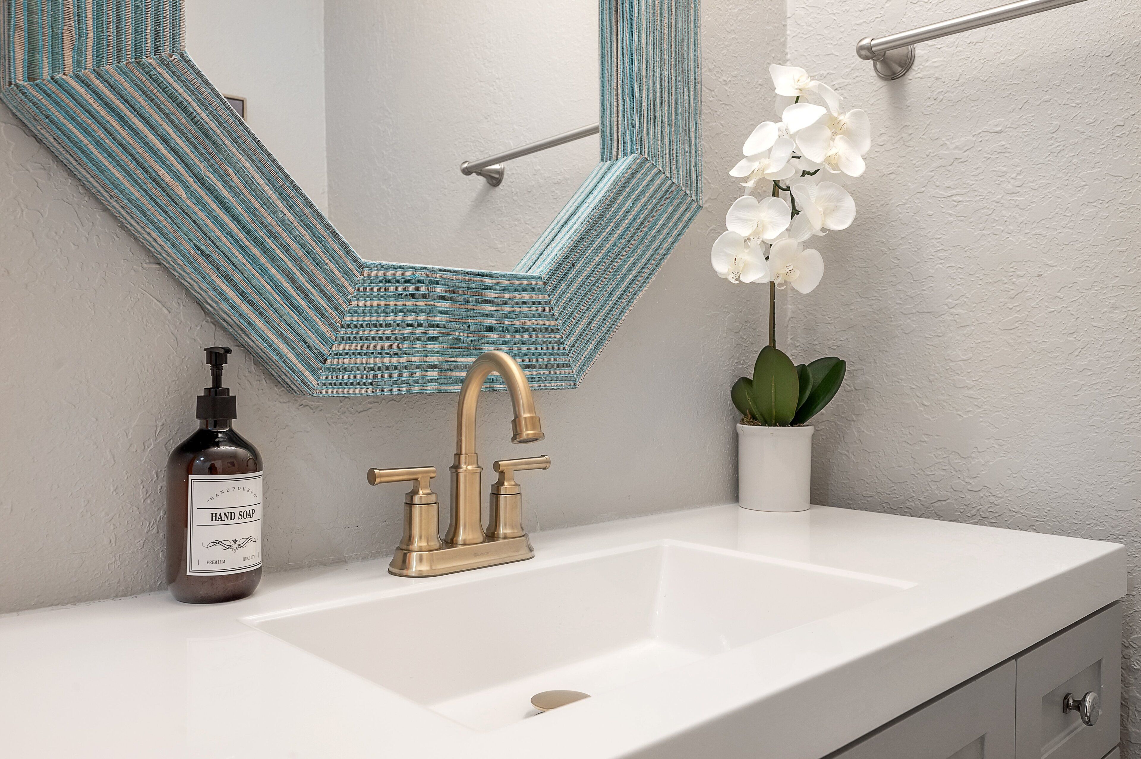 Une salle de bain avec un lavabo blanc, un robinet doré, un miroir à rayures, une orchidée en pot et un flacon de savon liquide.