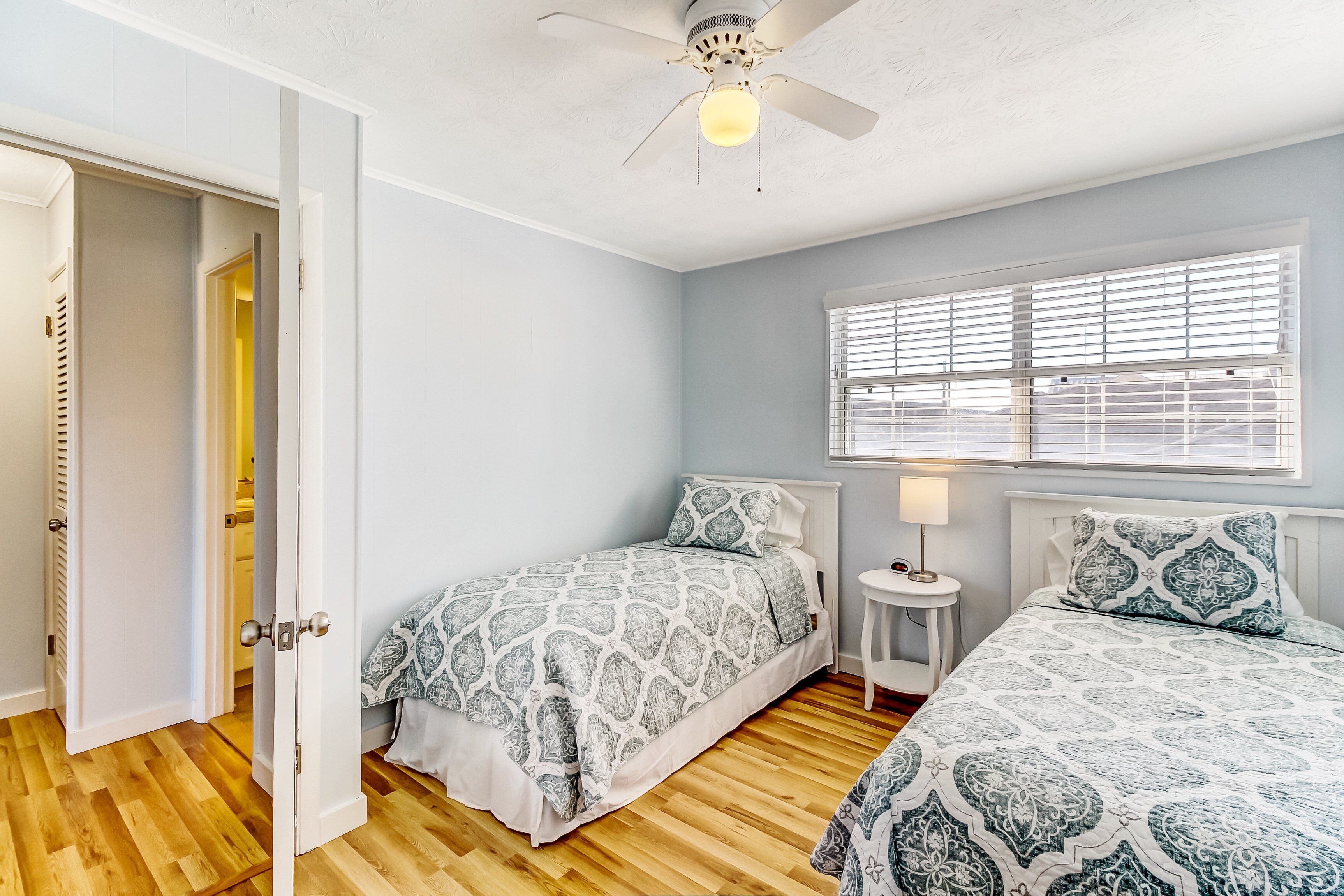 A bedroom with two beds, a ceiling fan, a window with blinds, and a small bedside table with a lamp.