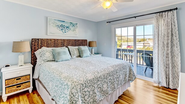 A bedroom with a bed, nightstands, a ceiling fan, a window with curtains, and a view of the outdoors.