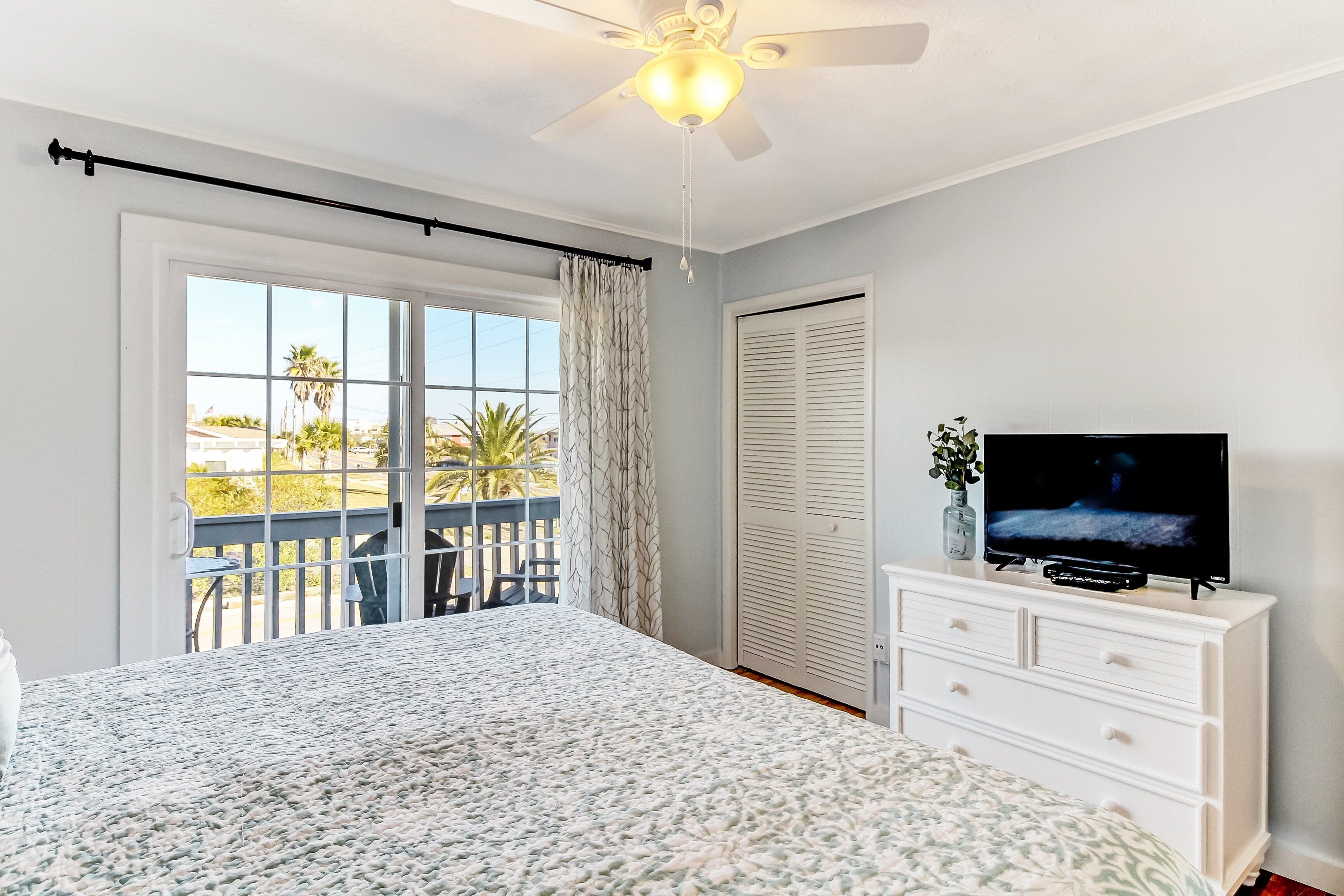 A bedroom with a bed, a television on a dresser, a ceiling fan, and a sliding glass door to a balcony.