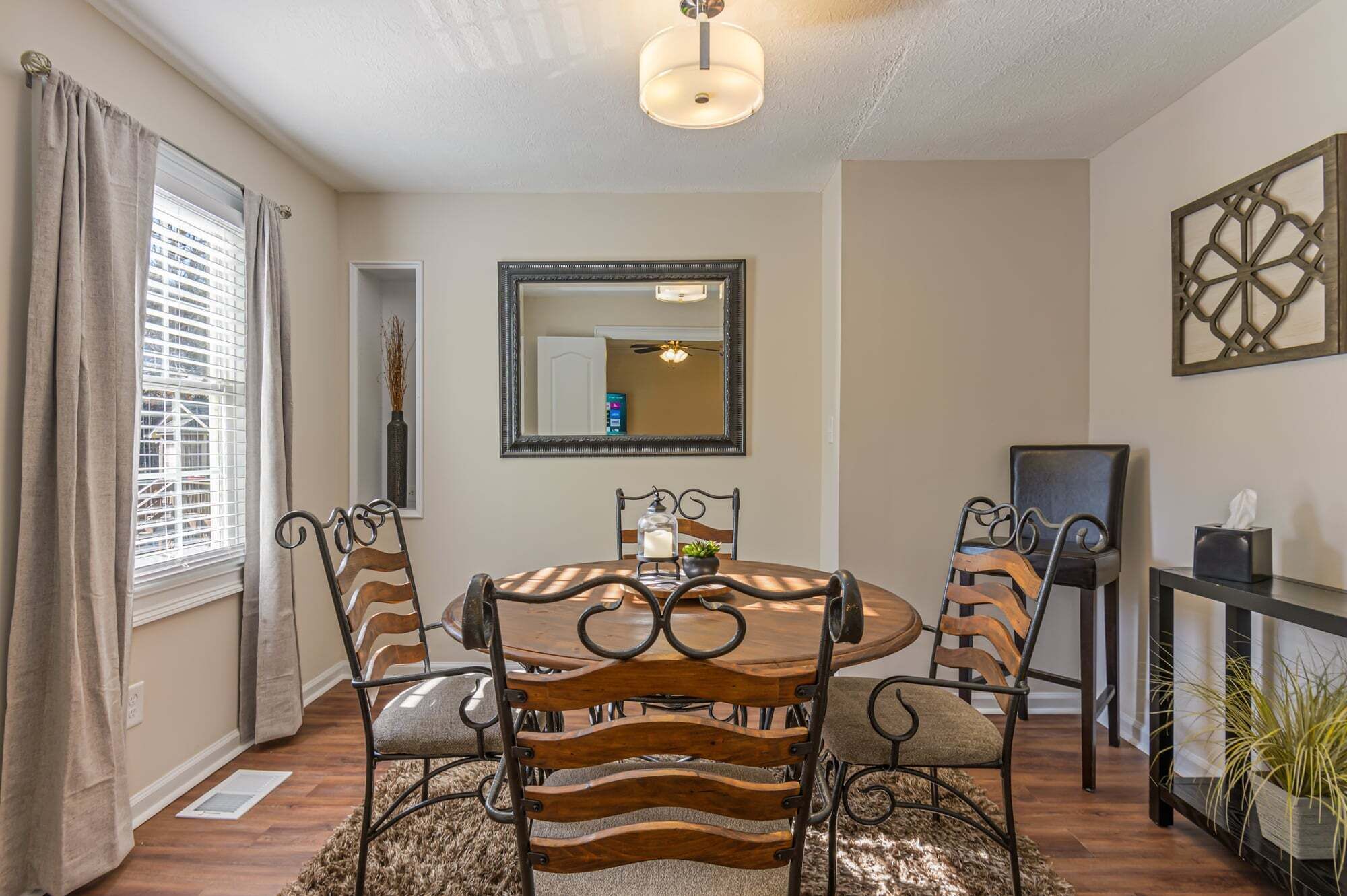 A dining area with a round table, chairs with wooden slats, a mirror, and a ceiling light fixture.