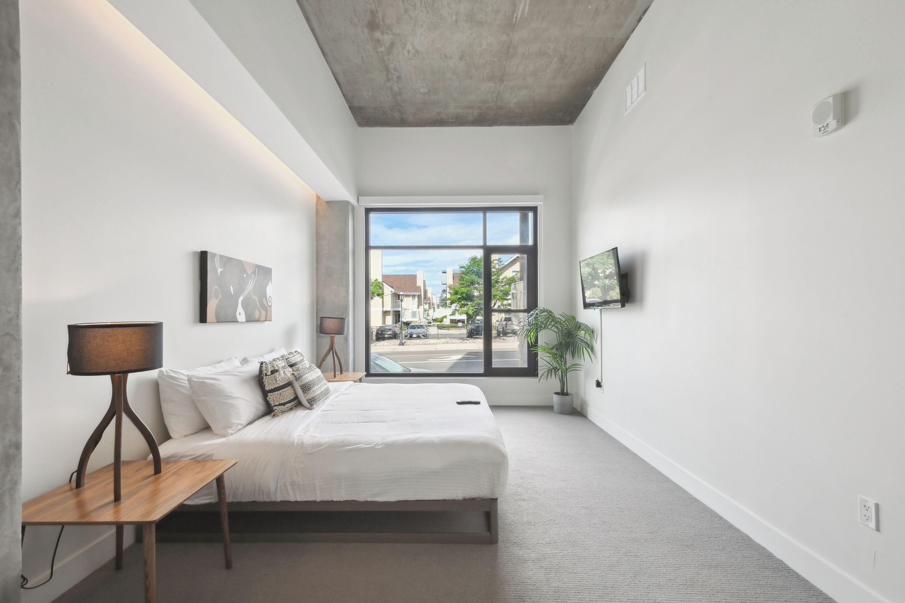 A modern bedroom with a large window, a bed, a bedside table, and a TV mounted on the wall.