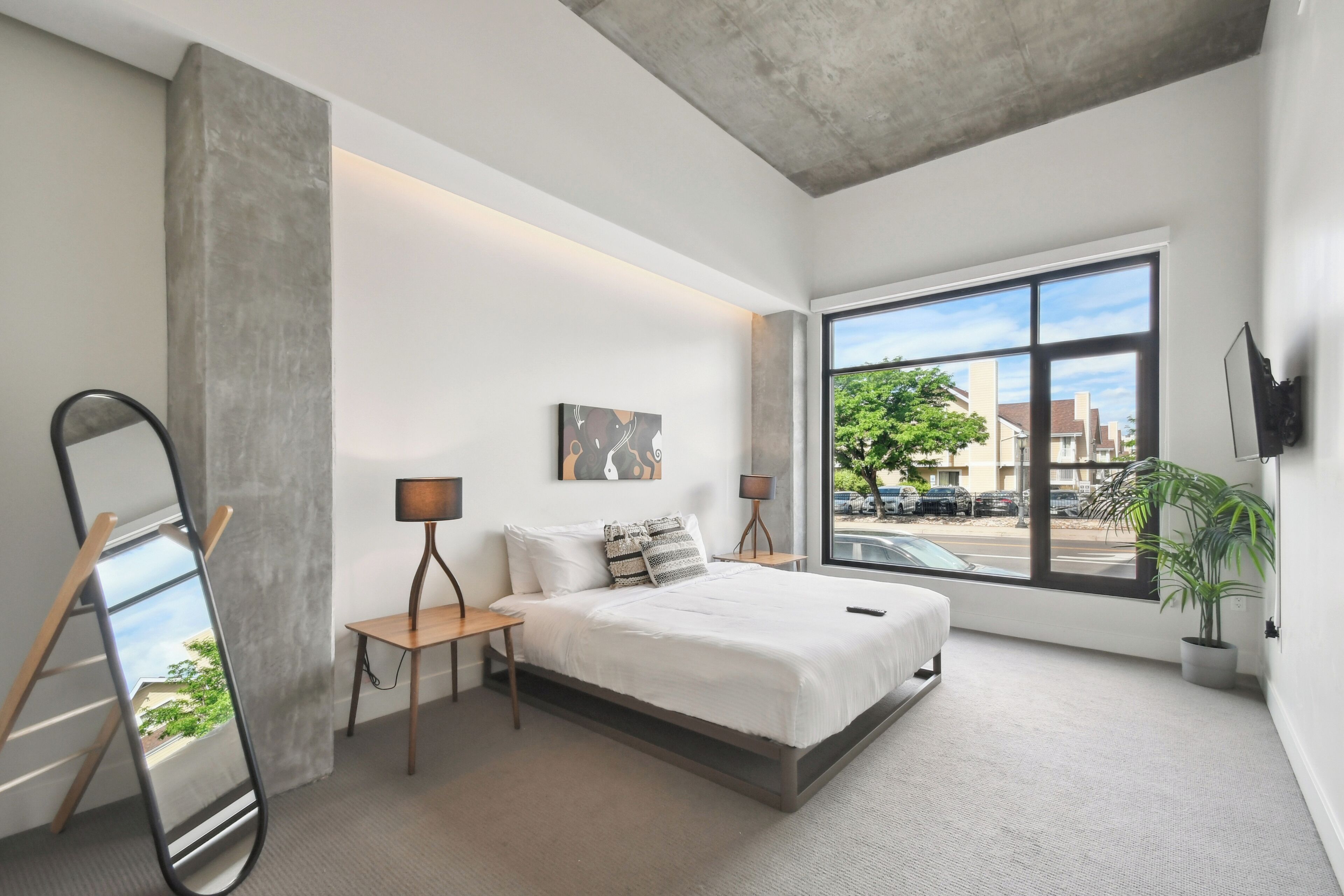 Un dormitorio moderno con un ventanal grande, una cama, mesitas de noche, un espejo y una planta en maceta.