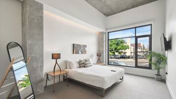 A modern bedroom with a large window, a bed, bedside tables, a mirror, and a potted plant.