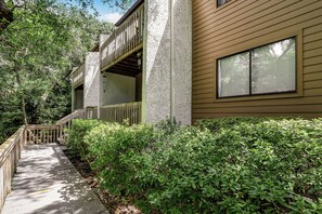 A wooden deck leading to a building with a balcony, surrounded by dense greenery. - 2745 Forest Ridge (Fernandina Beach)