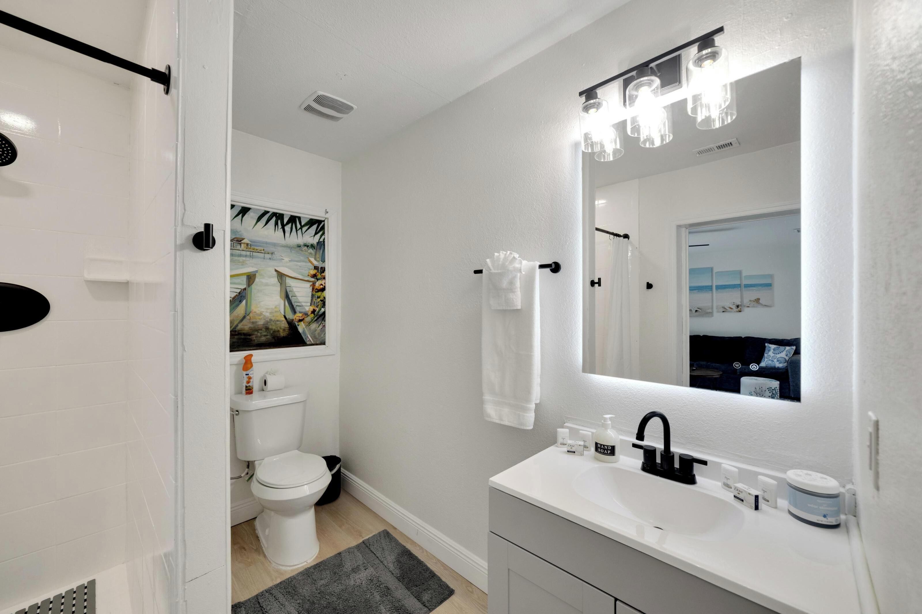 A modern bathroom with a white sink, black faucet, and a large mirror with lights. There is a toilet with a toilet paper holder and a picture on the wall.