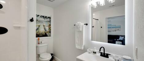 A modern bathroom with a white sink, black faucet, and a large mirror with lights. There is a toilet with a toilet paper holder and a picture on the wall.