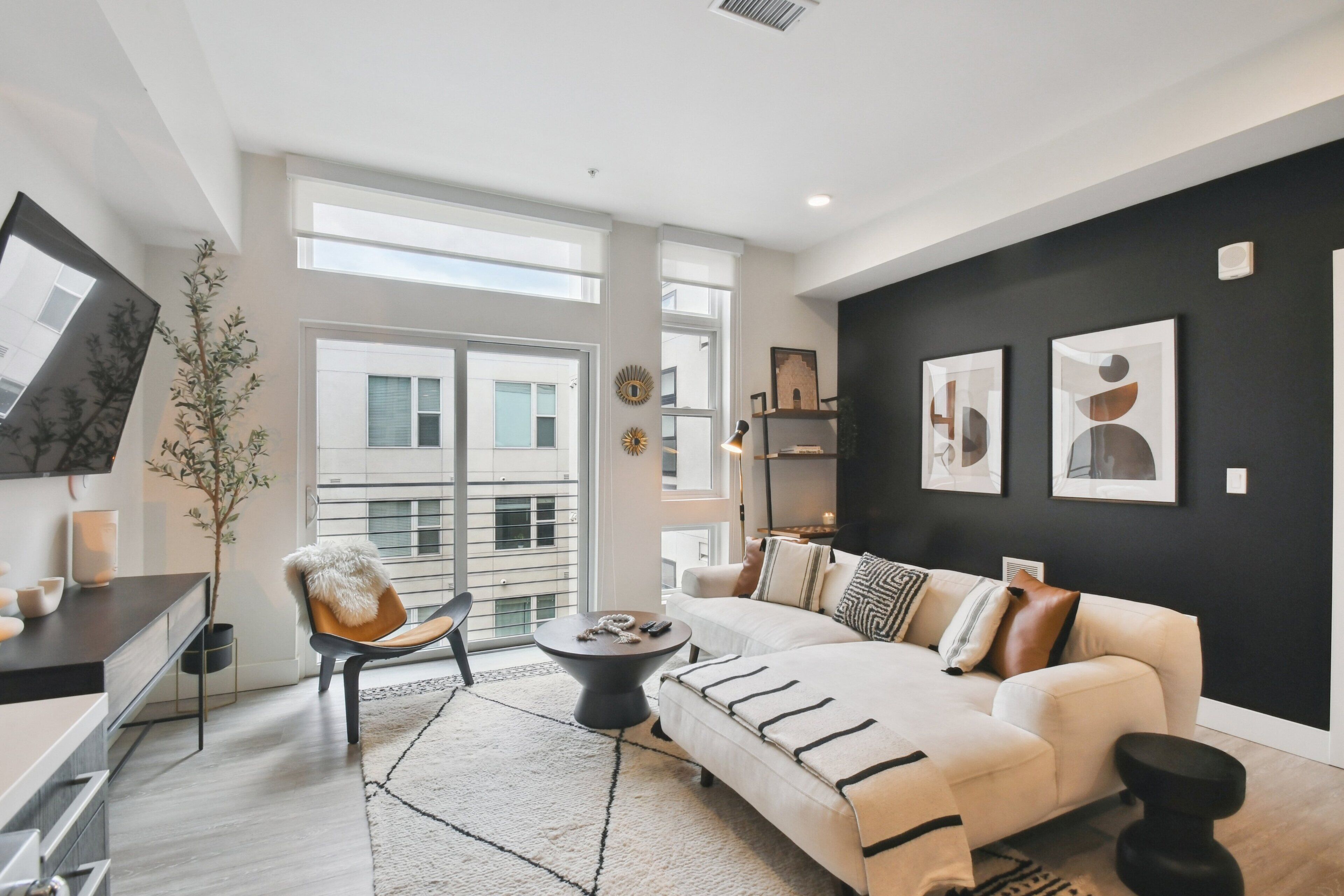 Un salon moderne avec un canapé, une table basse et un fauteuil. Une grande fenêtre laisse entrer la lumière naturelle et offre une vue sur l’extérieur.