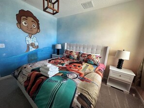 Une chambre d’hôtel avec un lit orné d’une couvre-lit aux motifs de personnages de dessins animés, une table de chevet avec une lampe et un mur décoré d’une fresque représentant des personnages de dessins animés.
