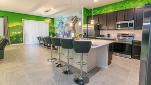 A modern kitchen with a central island, bar stools, stainless steel appliances, and a green mural on the wall. - 2476 Shanti Drive 12bed (Kissimmee)