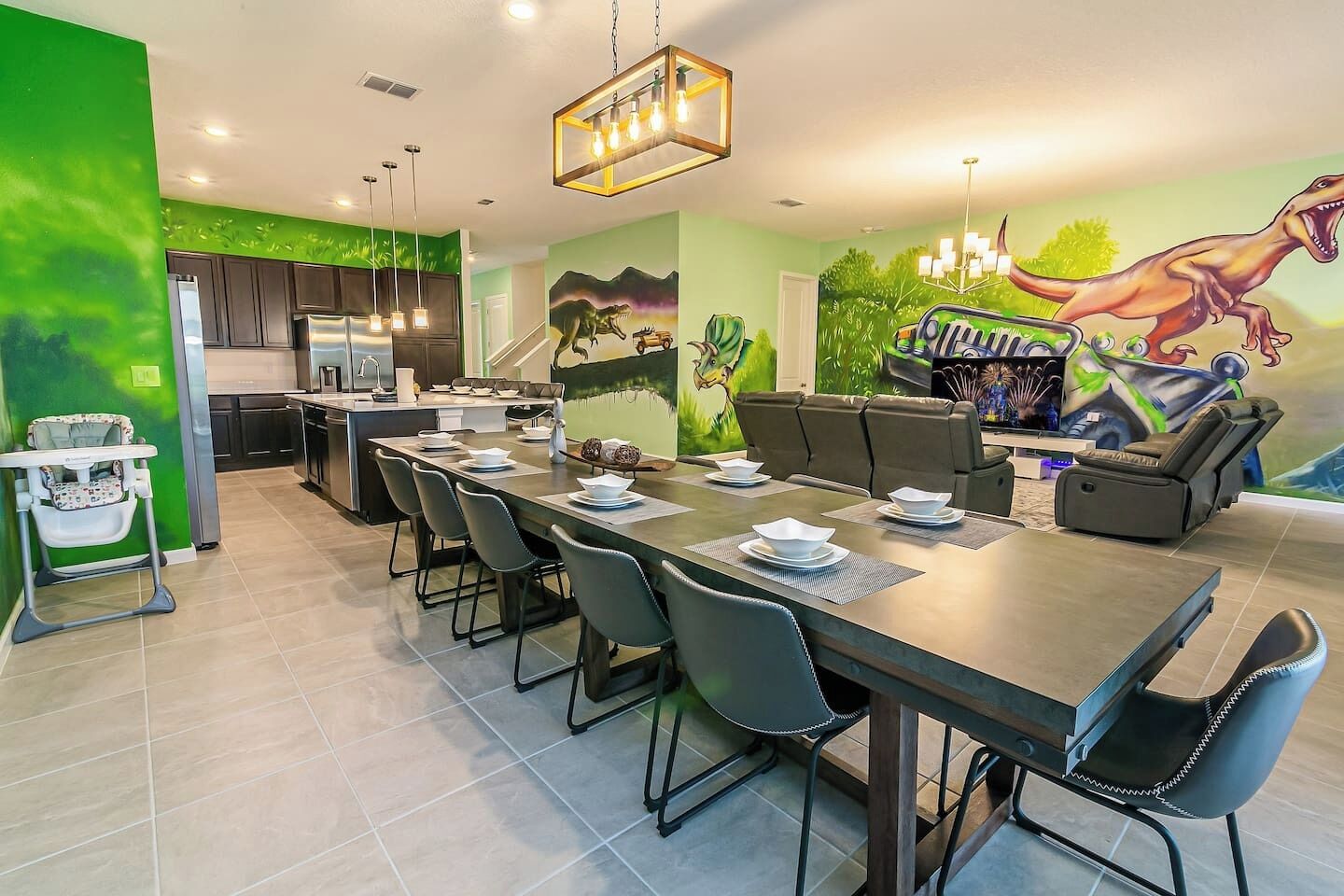 A modern kitchen with dark cabinets, a dining area with a long table and chairs, and a mural of dinosaurs on the wall.