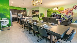 A modern kitchen with dark cabinets, a dining area with a long table and chairs, and a mural of dinosaurs on the wall. - 2476 Shanti Drive 12bed (Kissimmee)
