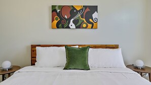 Una habitación de hotel con cabecera de madera, almohada verde, ropa de cama blanca y un cuadro abstracto y colorido en la pared.