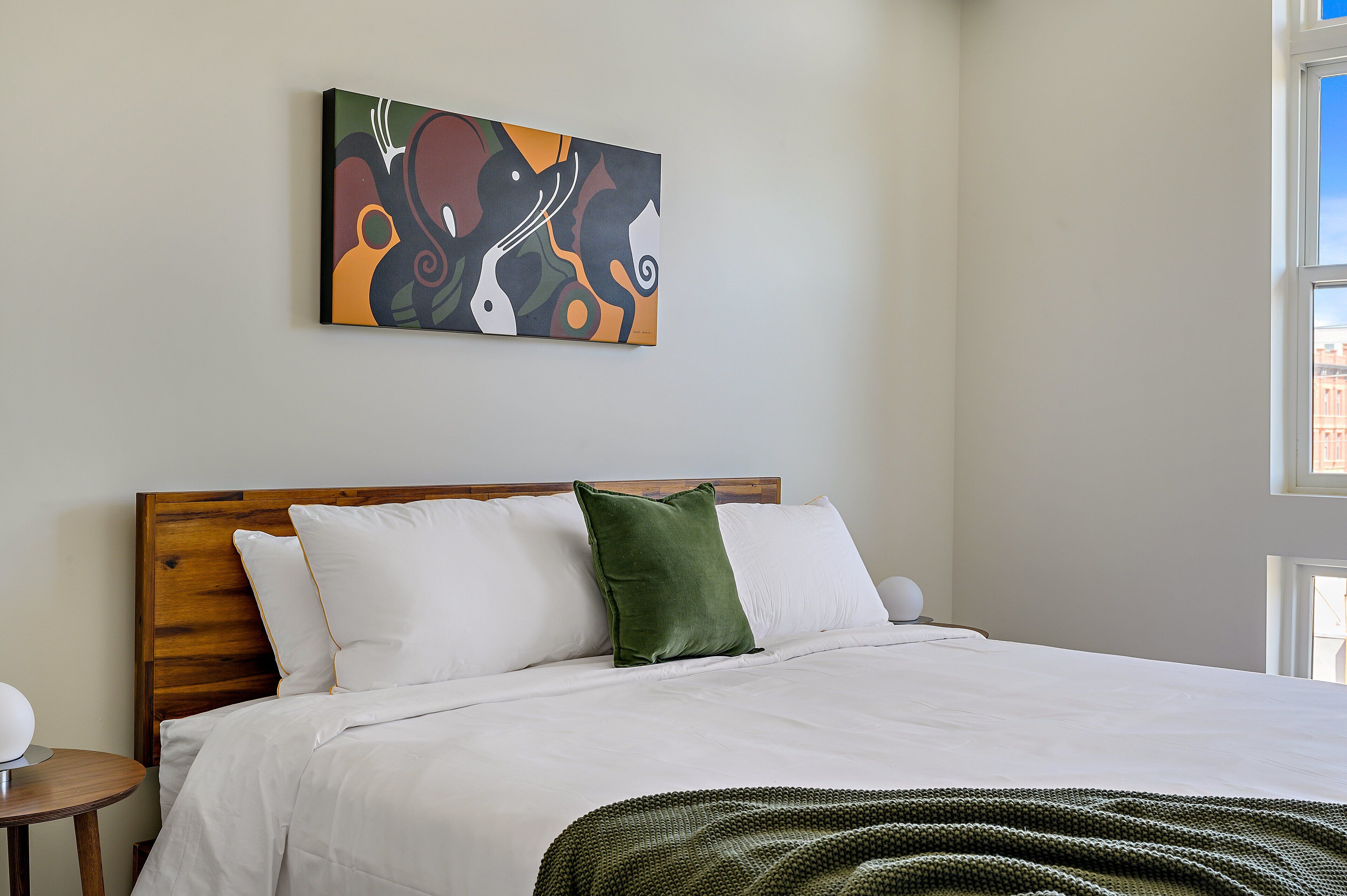 Ein ordentlich bezogenes Bett mit weißen Leinen und einer grünen Tagesdecke, ein Holz-Kopfteil und ein farbenfrohes, abstraktes Bild an der Wand.