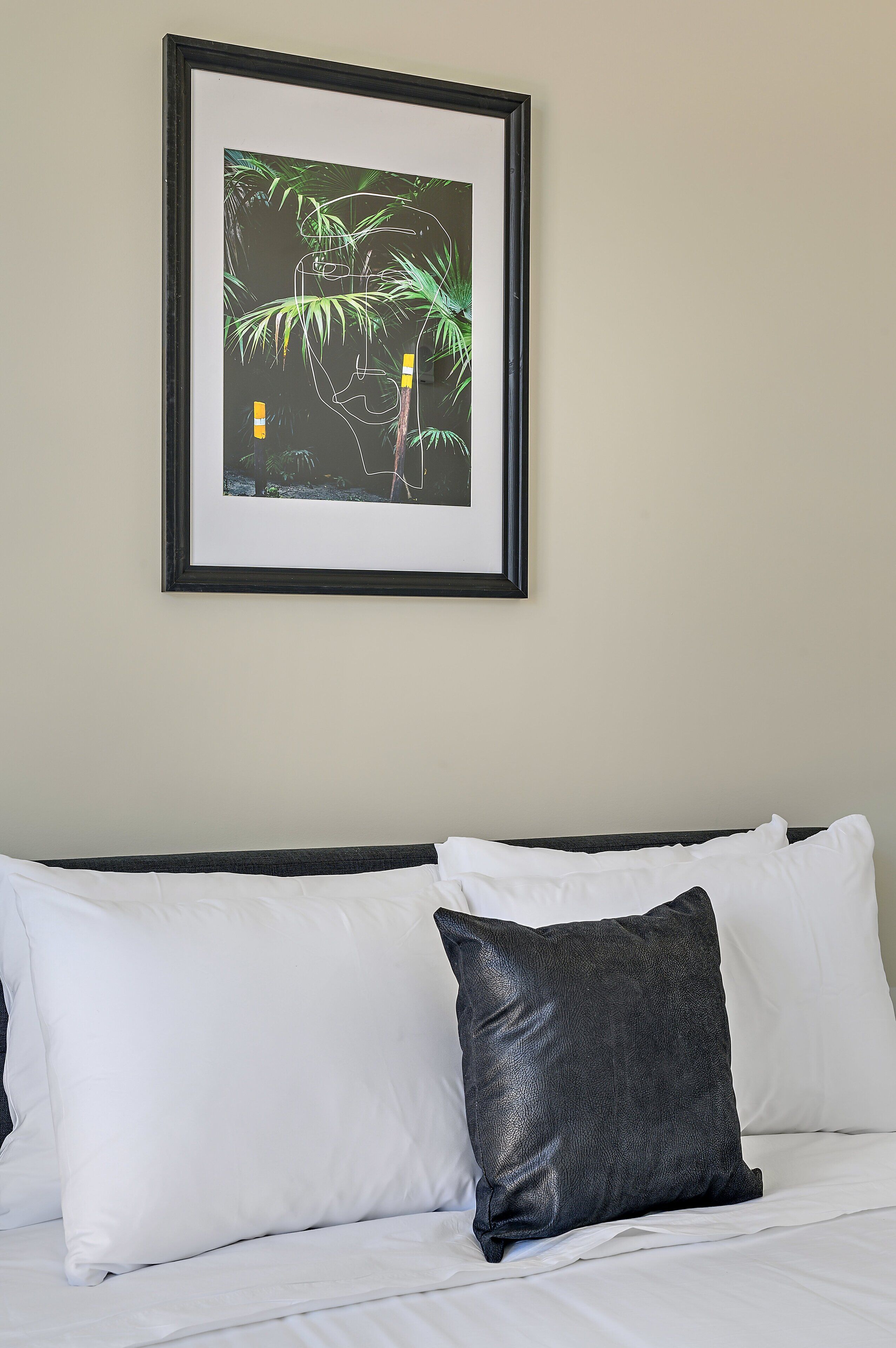 A bed with white and black pillows, a framed picture on the wall, and a plain wall.