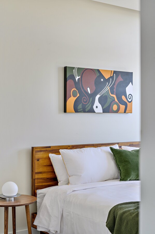 A neatly made bed with white bedding and a green pillow, a round table with a lamp, and a colorful abstract painting on the wall. - 2880: Canvas & Comfort at Espadin (Denver)